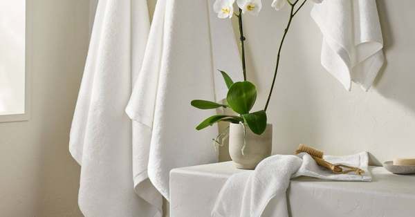 Stack of crisp white bath towels beside a marble tub with hanging robes and green orchid, creating a serene spa-like bathroom.