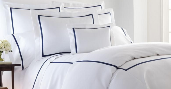 Elegant Strand bed with white cotton percale sheets and navy piped shams, duvet folded, sunlit shows long-staple weave and lush spa hotel luxury feel.